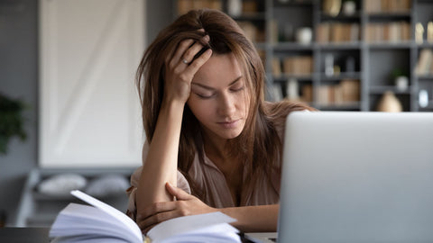 Supplements for Tiredness: Do They Work? A fatigued woman in front of a laptop and open notebook holding her hand to her head.