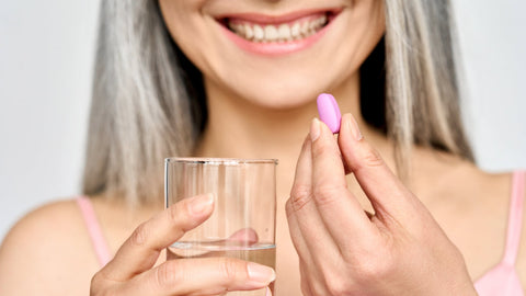 Best multivitamin for women over 70: A smiling woman in her seventies is holding up a multivitamin capsule and a glass of water.