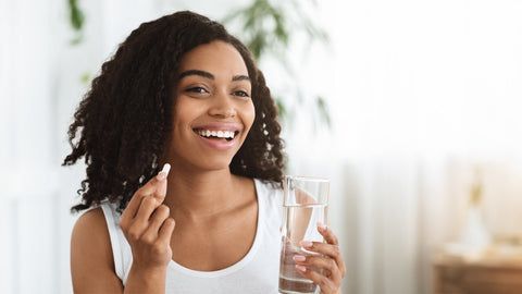 Smiling woman holding a supplement and a glass of water wondering when is the Best Time to Take Biotin (& 5 Reasons to Get Your Daily Dose) 