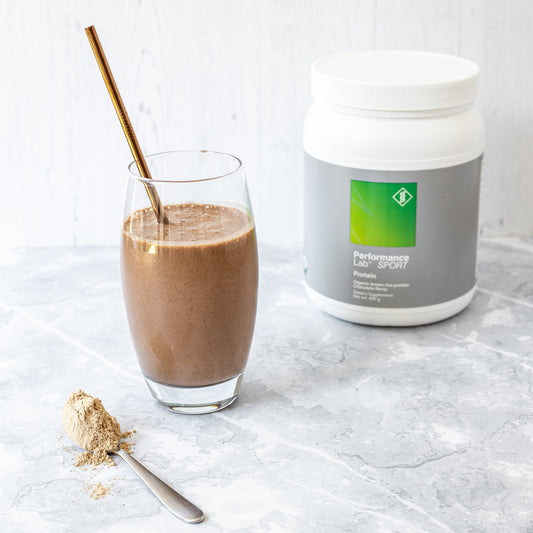 Glass with cacao protein smoothie and straw with protein tub in background