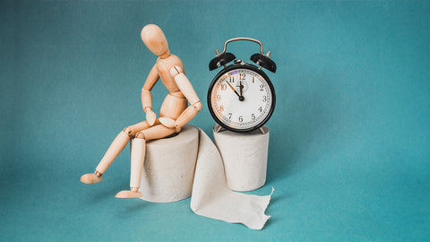  Do Iron Pills Cause Constipation? A wooden anatomical figure is sat on top of a toilet roll holding its stomach. Next to it sits a clock. Supporting the idea of constipation.