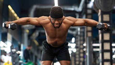 Man in gym with muscular shoulders and arms demonstrating a dumbbell rear delt fly movement.
