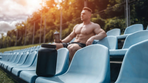 How Long Does Creatine Take to Work? Muscular man sitting outdoors in a sports park setting with a creatine shake in his hand and a bottle of creatine in the row of seats in the foreground.