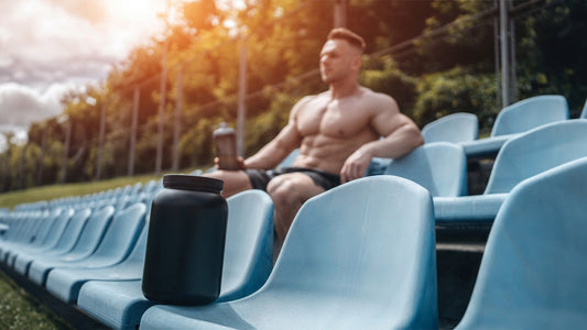 How Long Does Creatine Take to Work? Muscular man sitting outdoors in a sports park setting with a creatine shake in his hand and a bottle of creatine in the row of seats in the foreground.