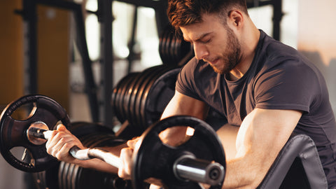 Man in his twenties with a focussed expression working on his biceps by lifting a barbell on a bench at the gym. He is focussed and wondering "How long should I spend at the gym?"