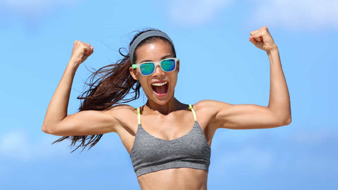 How to Get Skinny Arms: Female Guide to Slim, Thin, Toned Arms Fast. A toned arms female wearing sunglasses is shown flexing her biceps and smiling against a blue sky