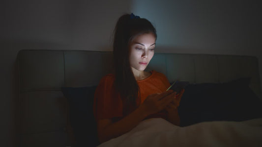 A dark haired woman sat up in bed in with the lights off. Her face is illuminated by the light of her phone. She is indulging in revenge bedtime procrastination.