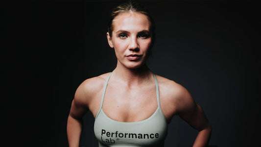 Low lighting Sophie Alisch wears a grey sports bra with Performance Lab across the front. She holds a strong stance and a neutral expression.