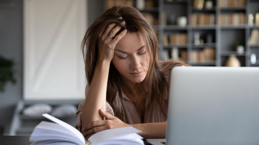 Supplements for Tiredness: Do They Work? A fatigued woman in front of a laptop and open notebook holding her hand to her head.