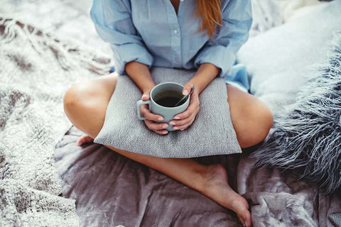 Is Coffee Bad For Gut Health? Woman in a blue shirt sitting cross legged on grey velvet blankets with a mug of coffee in her hands resting on a plush cushion.