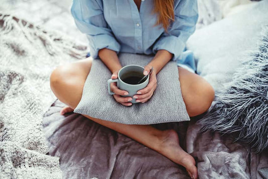 Is Coffee Bad For Gut Health? Woman in a blue shirt sitting cross legged on grey velvet blankets with a mug of coffee in her hands resting on a plush cushion.