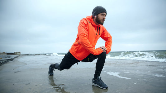 What Vitamins Should A 30 Year Old Man Take? - Top 7 Key Nutrients. A man in his 30s wearing an orange windcheater and hat is stretching on a windswept wintery shoreline.