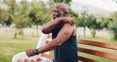 How to reduce joint inflammation quickly? Mature man in sports clothes sitting on a bench in nature holding his left muscular shoulder in pain.