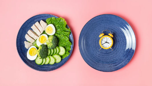 How many calories break a fast? An in-depth guide. A plate of lean protein (chicken, hard boiled eggs) and green vegetables (broccoli florets, sliced cucumber, lettuce) next to a plate with a clock on it.