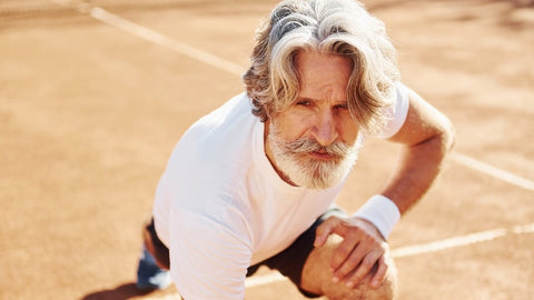 The best supplement for achy joints. An older man with gray hair and a beard kneels on an outdoor tennis court, wearing a white shirt and wristband.
